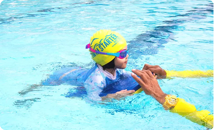 Child learning to swim with a yellow swimming board in the deep pool under the guidance of a Singapore swimming coach